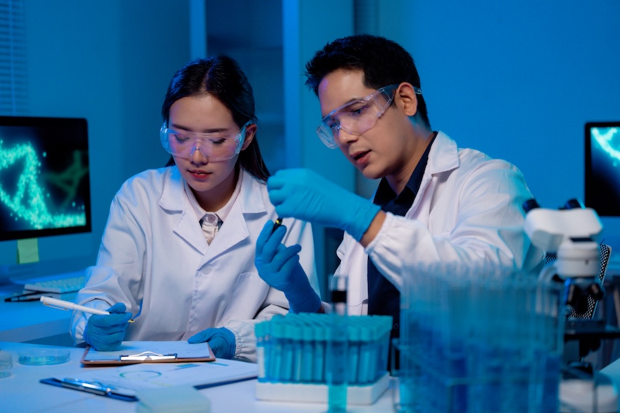 Two scientists wearing lab coats and gloves, collaborating on research involving test tubes and taking notes on a clipboard in a modern laboratory