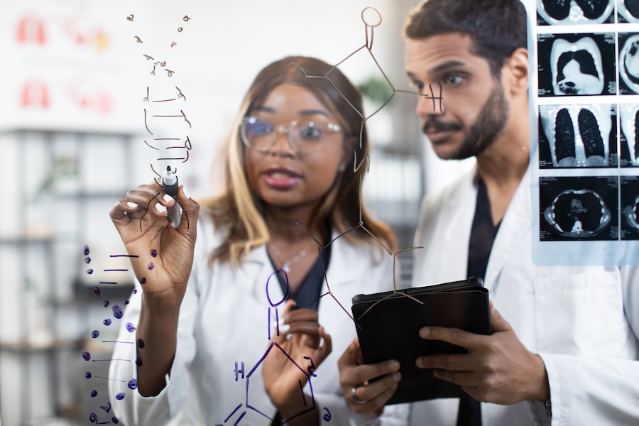 Close up portrait of two diverse scientists doctors chemists, standing together near glass wall at bright office and writing DNA structure. Focus on hand of afro woman writing on the wall